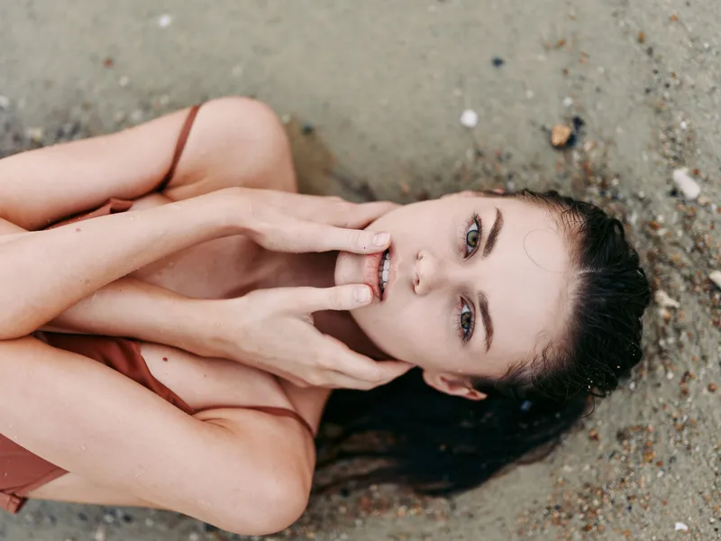 Portrait of a relaxed woman lying on sandy ground, head tilted, hands framing her quiet face, soft light, warm tones, subtle mood, intimate beach moment.