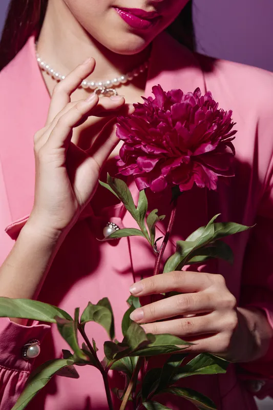 Close-up of a stylish woman in a pink outfit holding a vibrant burgundy flower and adjusting a pearl necklace. Fashion, beauty, creative concept with elegant details.