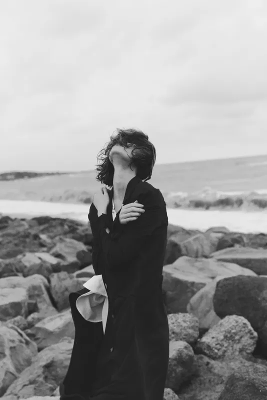 Monochrome scene of a woman on a rocky shoreline, wind in the hair, hands raised to shield the face, moody and introspective, highlighting resilience against the stormy sea backdrop