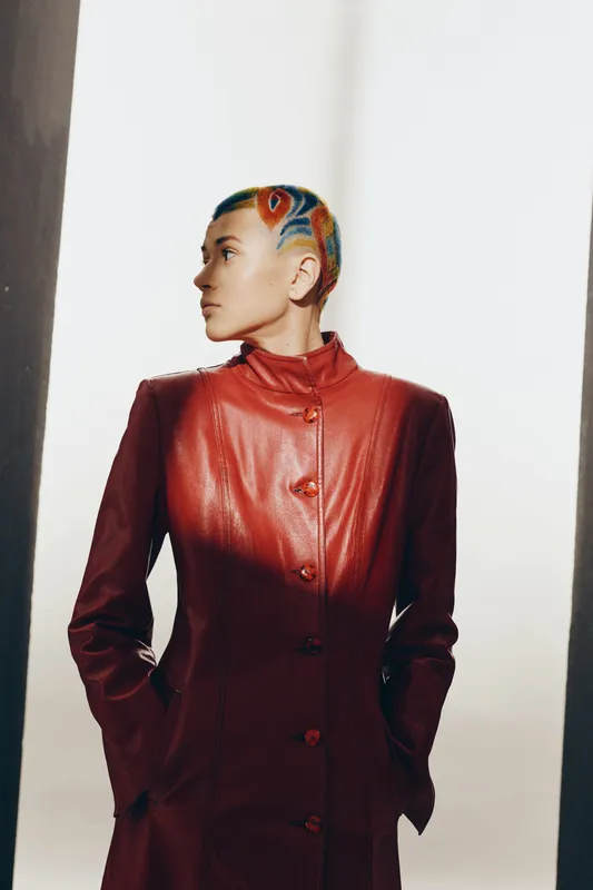 Young woman with colorful short hair wearing red leather coat stands confidently against bright background, showcasing edgy fashion and urban style attitude.