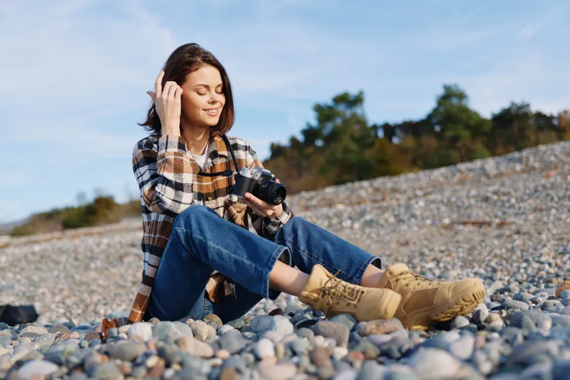 Woman sits on a pebble beach in casual outfit, headphones on, holding a camera and enjoying outdoor scenery, relaxation and focus in nature with bright light.