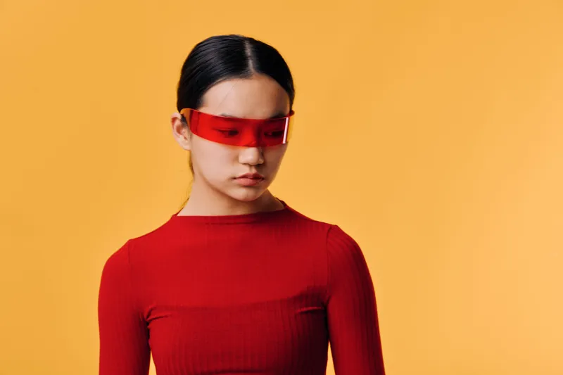 Young woman in red top posing with futuristic red visor on yellow background. Portrait of stylish girl with straight hair looking down, modern fashion and minimal concept in studio.