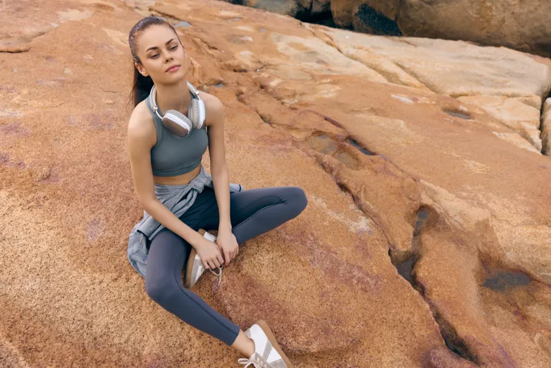 Young woman in activewear sitting on rocks, wearing headphones, reflecting calm and determination in nature with earthy tones, ideal for fitness and wellness themes