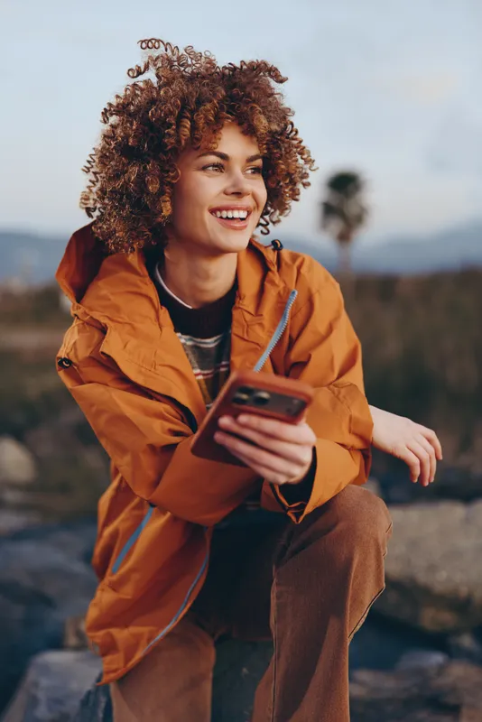 Woman with curly hair smiling outdoors, wearing orange jacket, using a smartphone in nature. Soft daylight and blurred mountains create relaxed lifestyle mood for travel and communication.