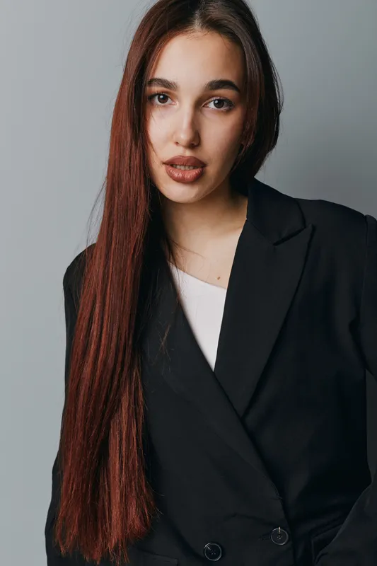 Young woman with long brown hair wearing black blazer and white top standing against gray background looking at camera with confident expression in studio portrait.