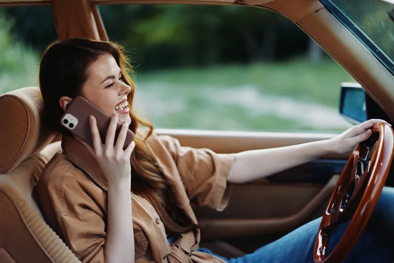 Young woman smiling while talking on smartphone in a vintage car, conveying happiness and relaxation in nature, wearing casual beige shirt, with green background.