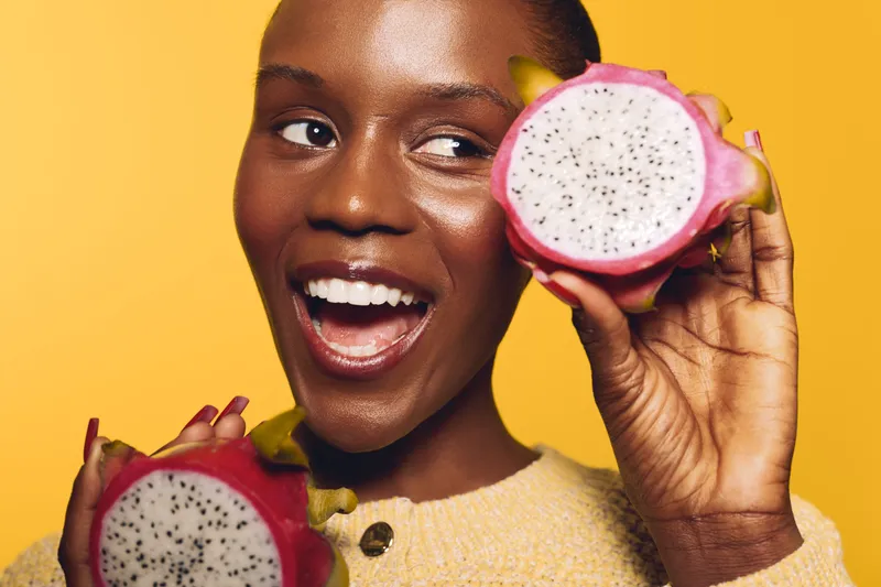 Happy African woman with dark skin holding two halves of dragon fruit near face on yellow background. Concept of healthy lifestyle, tropical fruit, nutrition and natural beauty.