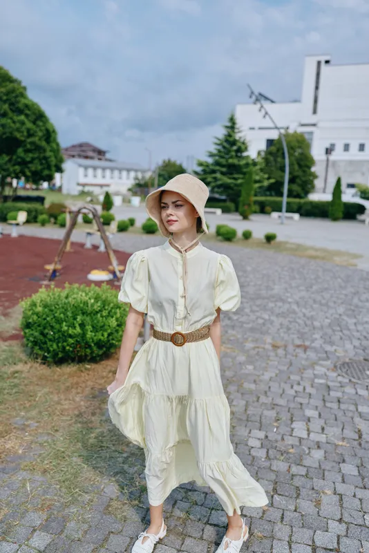 Woman wearing a light cream dress and straw hat strolls along a cobblestone path in a sunny outdoor setting, exuding relaxed summer style.