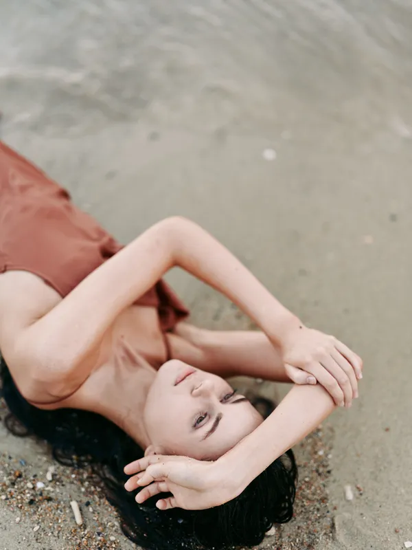 A woman lies on a sandy beach with arms raised above her head in a relaxed sunlit pose. Soft tones and gentle breeze create a calm, contemplative summer atmosphere.