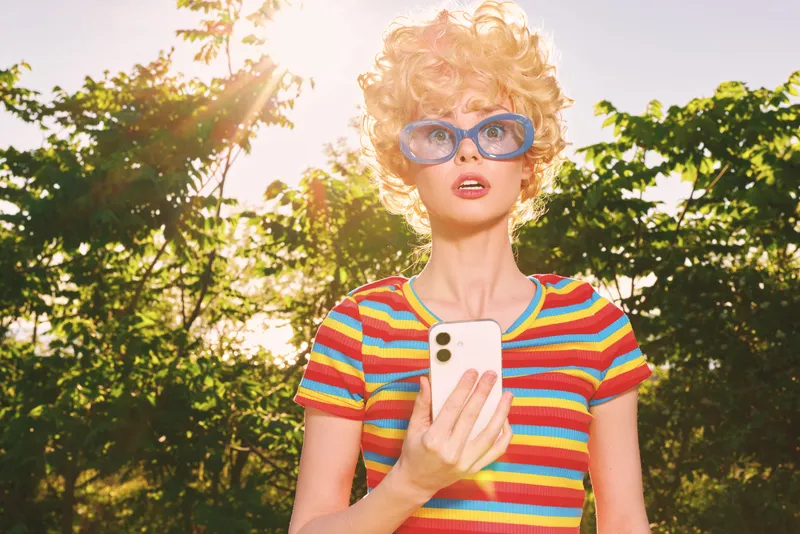 Young woman with curly blonde hair in a colorful striped tee stands outdoors under bright sun, holding a smartphone and wearing bold blue sunglasses, conveying playful confidence.