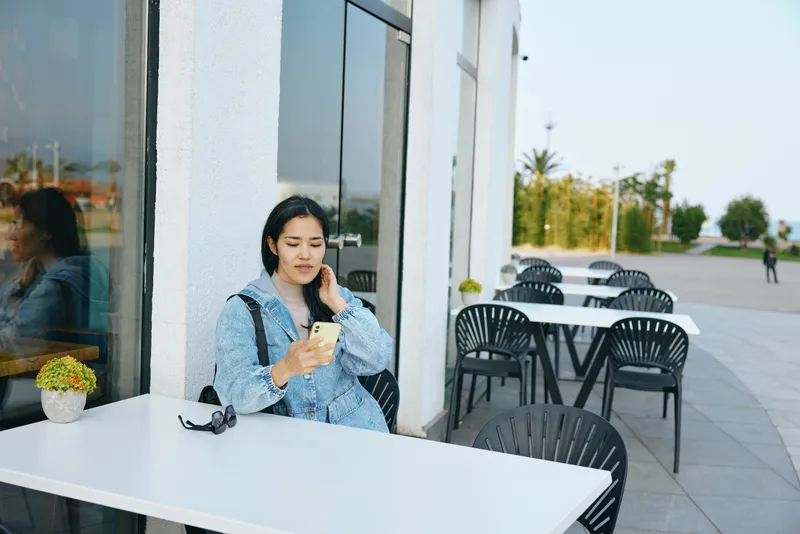 Woman sitting at outdoor cafe, browsing on phone beside a white table, sunglasses on the edge, denim jacket, calm urban mood, sunny day, casual lifestyle scene perfect for stock and social media
