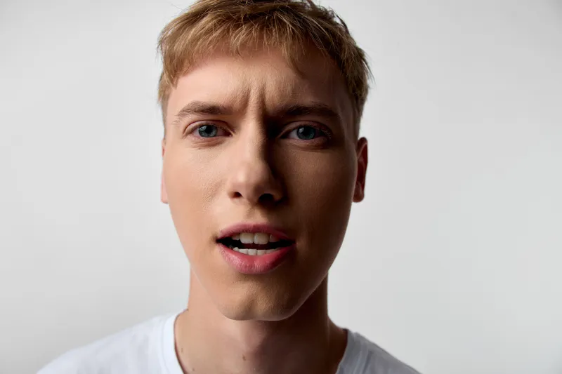 Close up of a young man with surprised expression, open mouth and wide eyes, studio lighting, white shirt, sharp detail, clean background, candid and expressive mood