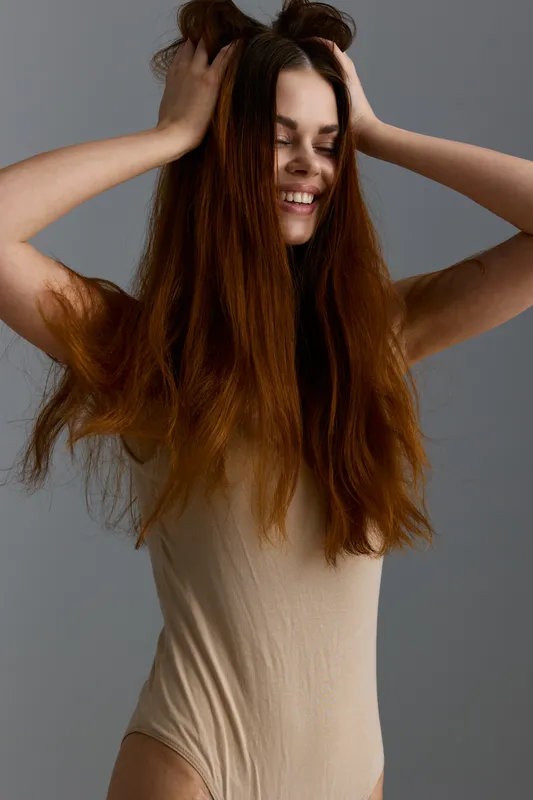 smiling woman with long brown hair wearing beige bodysuit, expressing joy and carefree mood against a gray background in soft lighting, natural beauty concept.
