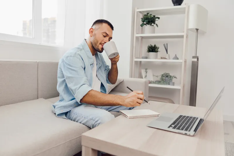 Focused man enjoying morning coffee while writing in a notebook, surrounded by a cozy, modern living space with natural light and minimal decor.