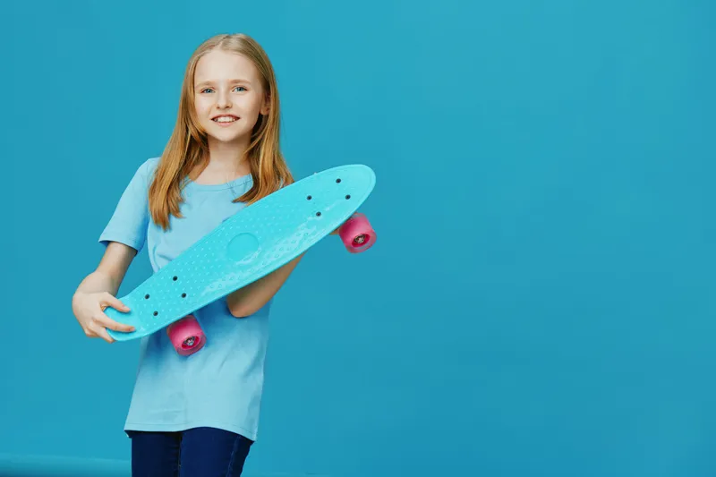 Cheerful girl with long hair holding a blue skateboard against a vibrant blue background, showcasing youthfulness and fun in summer fashion