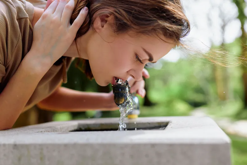Woman drinking water from public fountain in park, closeup portrait of young female leaning over stone basin, natural light, refreshing moment and outdoor hydration.