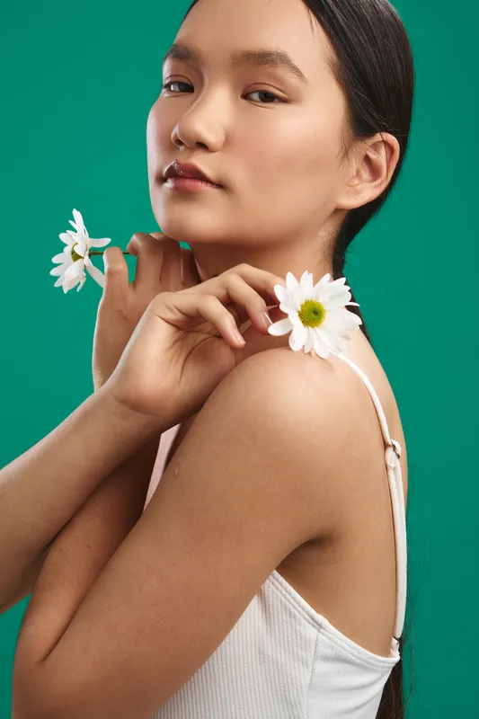 Asian woman poses with white flowers against a solid green backdrop, soft studio lighting, minimal wardrobe, conveying calm elegance, natural beauty, and a serene, artistic mood.