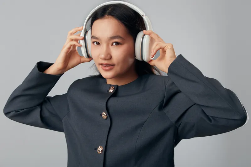 Young woman wearing large headphones adjusting them with both hands, smiling and looking at camera, studio portrait with gray background in casual dark clothing outfit.