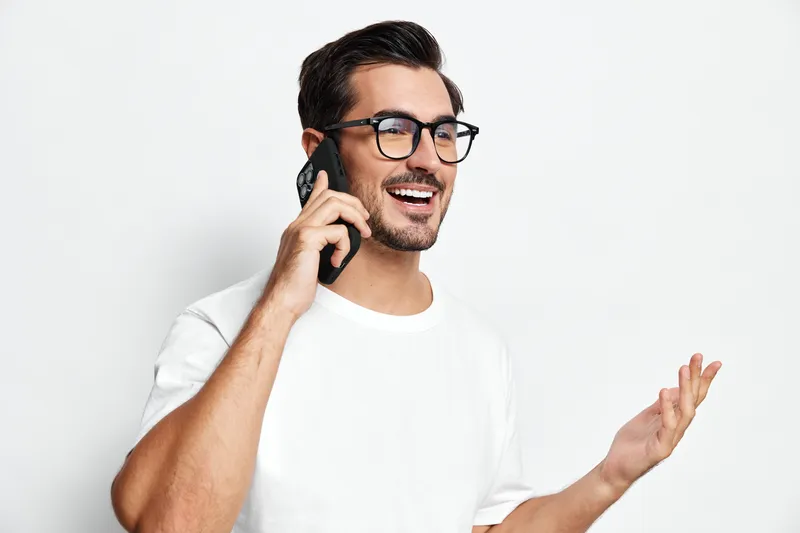 Smiling man wearing glasses talking on mobile phone, expressing joy and engagement while standing against a clean, minimalist white background. People lifestyle concept