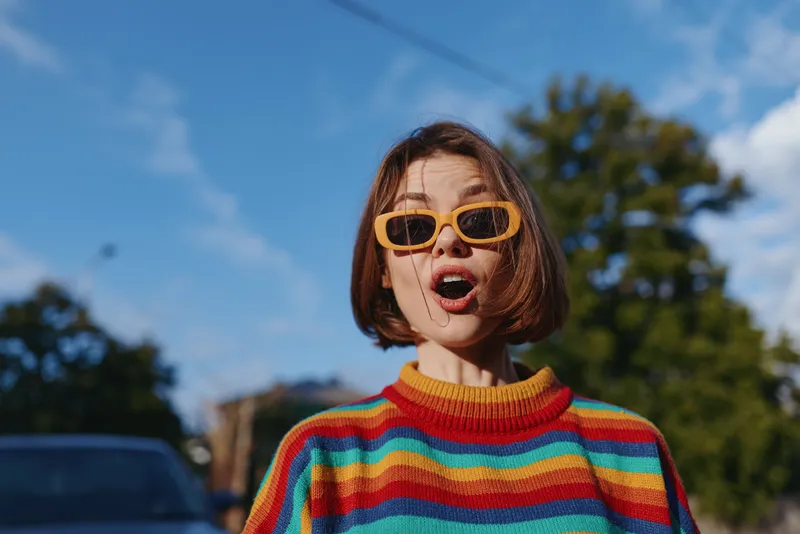 Woman young adult striped sweater yellow sunglasses surprised expression outdoors on sunny street, casual outfit with bob haircut, playful travel lifestyle portrait with open mouth.