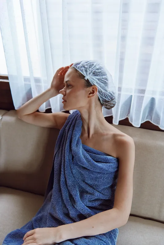 woman relaxing wrapped in towel and hair cap sitting on sofa near window with white curtains, natural light, calm indoor atmosphere