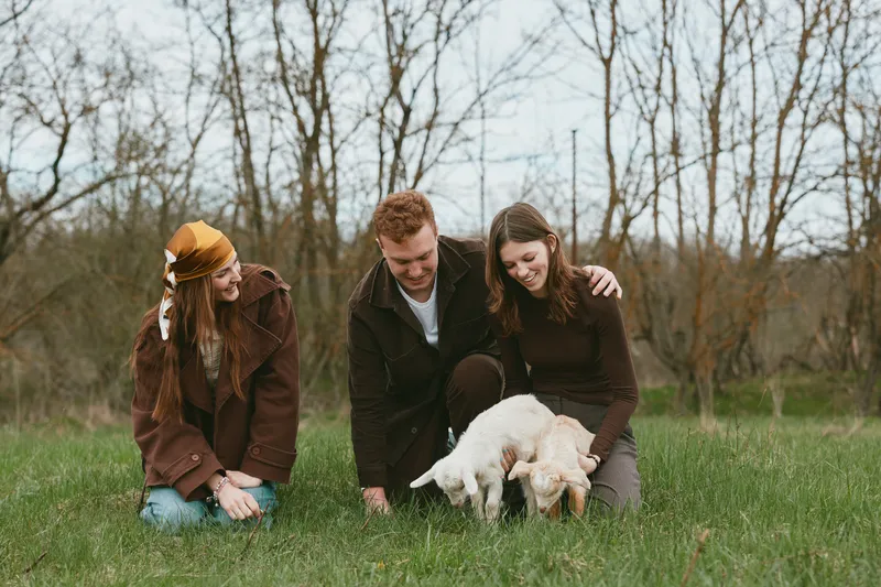 Lifestyle moment with friends outdoors, goats and kids in a sunlit countryside, together and smiling, capturing relaxed companionship, warm tones, and a playful, wholesome mood in a natural setting.