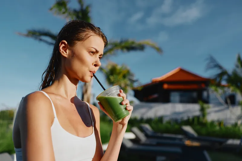 Smiling woman with wet hair enjoys a green smoothie by a poolside resort, highlighting healthy lifestyle, self care, hydration, and outdoor relaxation in bright daylight.