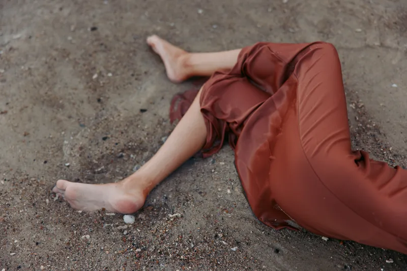 Close view of a person lying on dusty ground, wearing a flowing reddish dress, bare feet, soft fabric folds, and textured gravel creating a quiet, intimate mood.