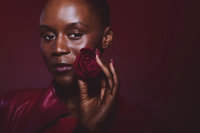 Portrait of African American woman with dark skin holding red rose near face. She wears red leather clothing and dark red nail polish. Concept of beauty, elegance, and floral aesthetics.