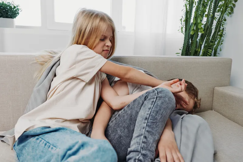 Two young children are fighting aggressively on a sofa in a bright living room, showing conflict and sibling rivalry.