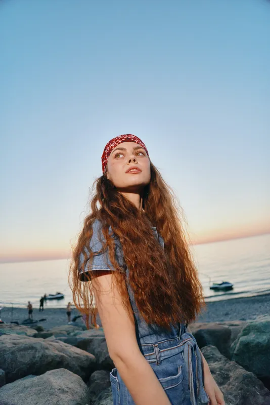 Lifestyle woman in modern boho-western denim street style with long hair and headband, standing on rocky beach at sunset in warm film color tones.