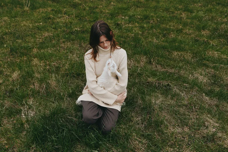 A young woman in a beige sweater gently holds a white baby goat while kneeling on green grass in a natural outdoor setting during a calm lifestyle moment.
