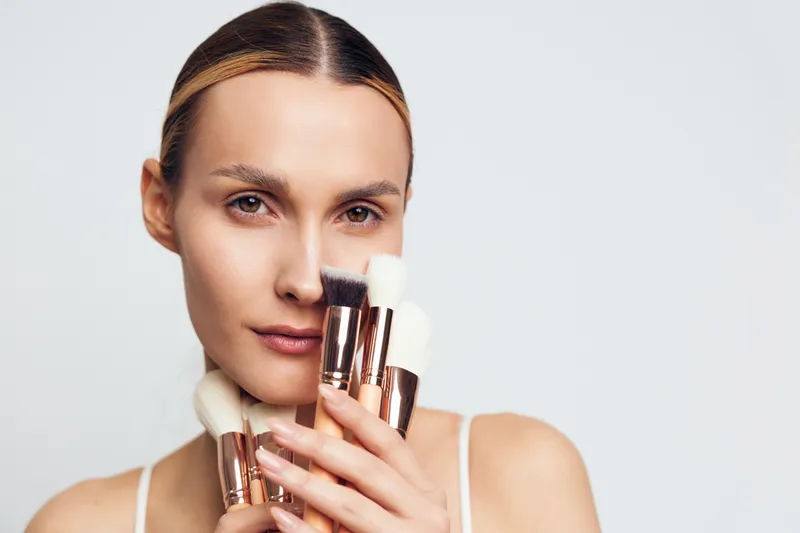 Young woman with clear skin holding makeup brushes close to her face, neutral expression, soft lighting, minimal makeup, plain background, beauty concept