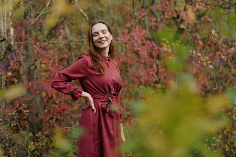 Genuine portrait of a smiling woman in a red dress standing among autumn foliage, capturing authenticity through relaxed pose, warm light, and natural mood.