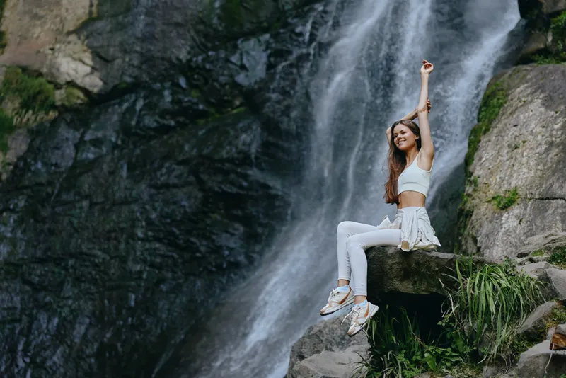 young woman sitting joyfully on rock near waterfall wearing white activewear and sneakers, surrounded by lush green foliage and natural scenery outdoors