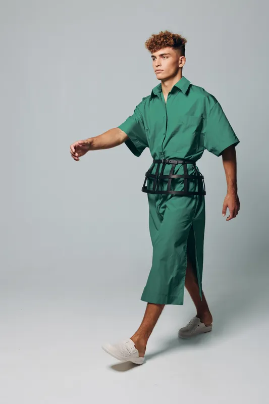 Young man walking in studio wears green oversized shirt dress with black waistbelt and thumbs up white slip on shoes standing on gray background active movement casual fashion look