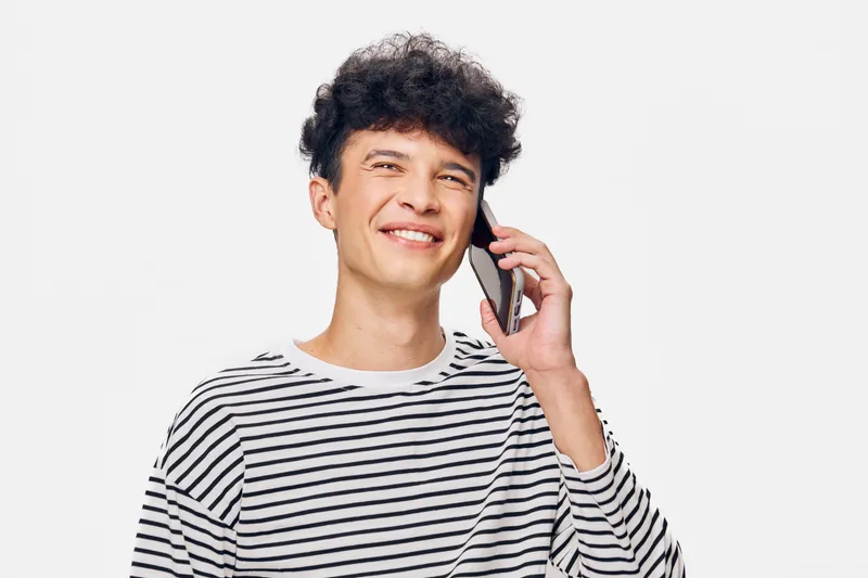 young man smiling talking on smartphone wearing striped shirt with curly hair on white background, casual style with cheerful expression