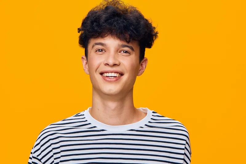 smiling young man with curly hair wearing striped shirt on bright orange background, cheerful expression showing happiness and youthfulness.