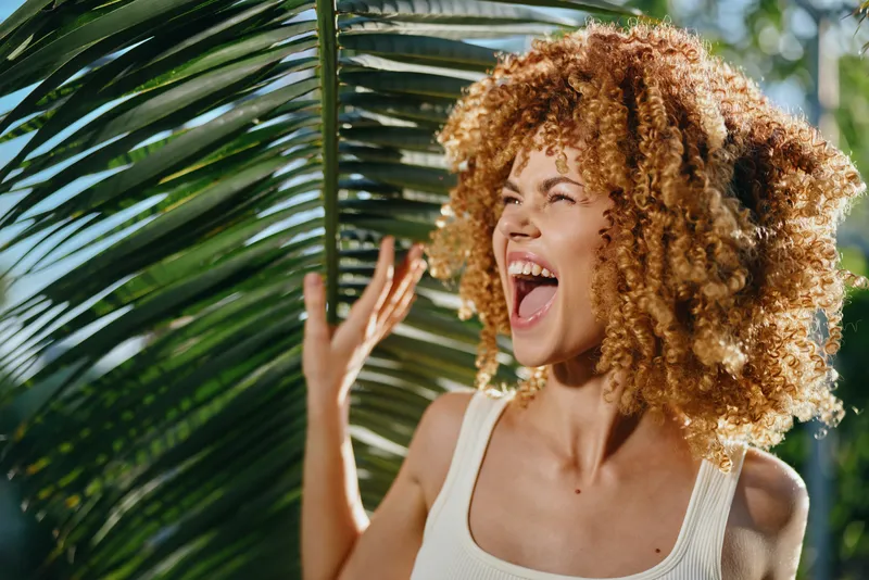 cheerful woman with curly hair laughing outdoors near palm leaves in bright sunny weather, wearing light casual tank top, natural summer vibe