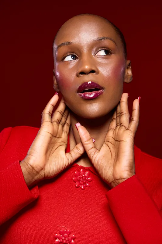 Portrait of a confident black woman with dark skin wearing a red sweater, glossy lips, and manicured nails. She poses with hands near her face against a red background, fashion concept.
