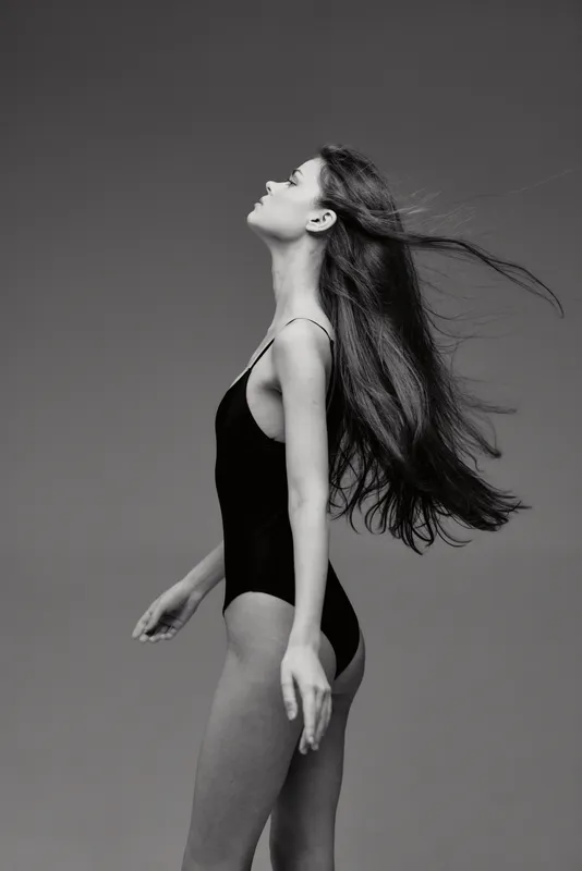 Black and white profile of a young woman with long hair in a black bodysuit against simple background.