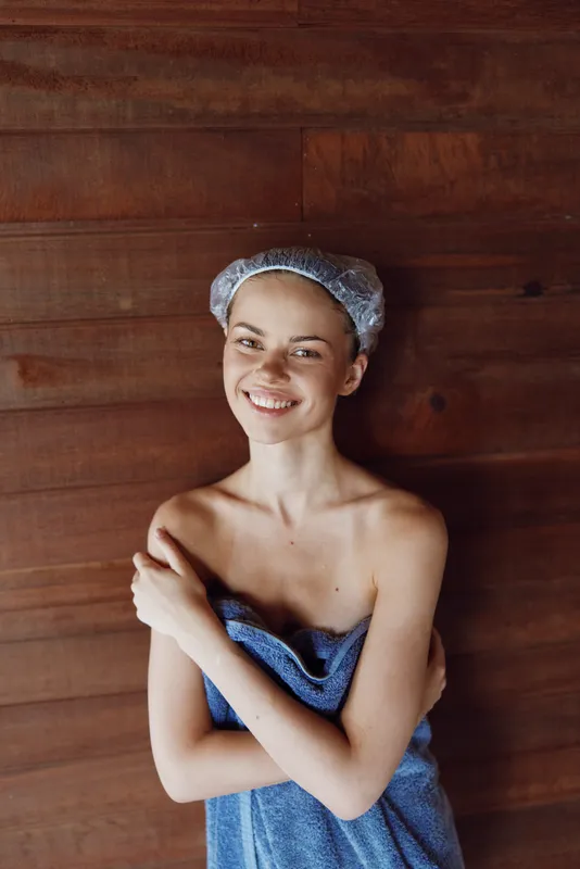 smiling woman wrapped in blue towel with shower cap on head standing against wooden wall, natural light, relaxed mood, skincare and wellness concept