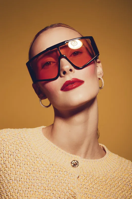 Stylish woman with short hair wearing oversized orange sunglasses and red lipstick poses against warm brown background. Fashion portrait with modern accessories and confident expression.
