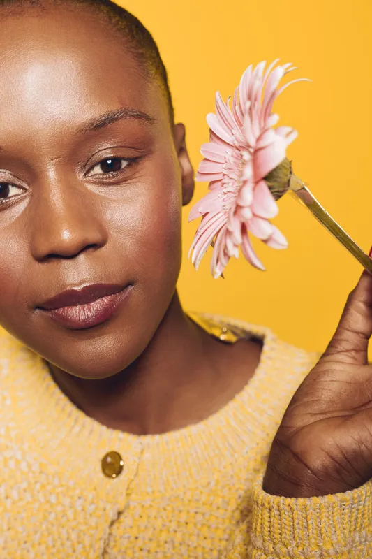 Close-up of a smiling black woman with dark skin holding a pink flower near her face against a yellow background. Concept of natural beauty, skincare, and floral wellness.