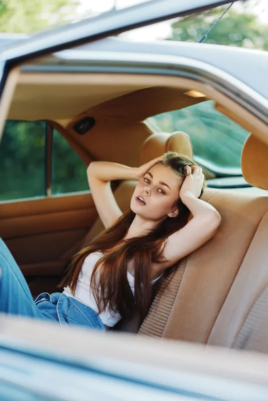 Young woman relaxing in a car, showcasing casual fashion, flowing hair, and natural beauty in a lush green outdoor setting.