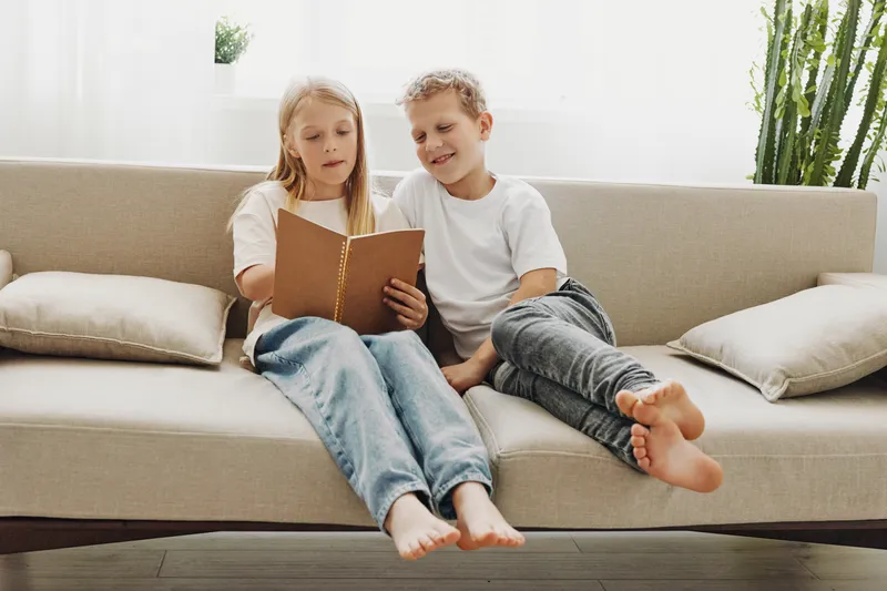 Two children sitting barefoot on a beige sofa reading a book together in a bright room with natural light and plants. Concept of childhood learning, friendship, and cozy home atmosphere.