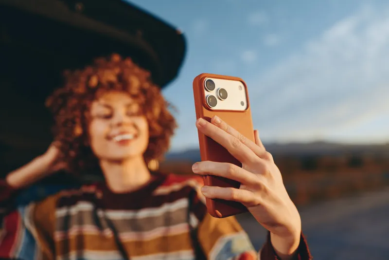 Woman lifestyle selfie smartphone in hand, curly hair smiling, outdoors in golden hour light, casual street setting, shallow depth of field blur, relaxed friendly mood at sunset