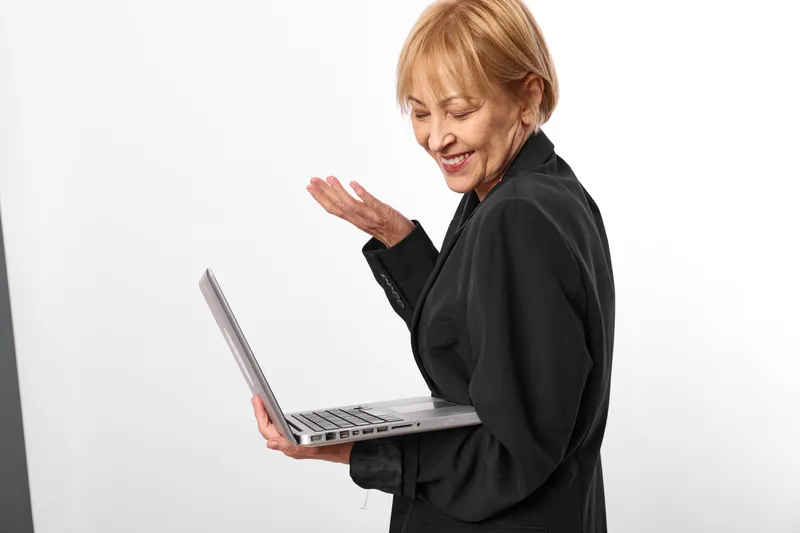 Smiling senior woman with laptop, one hand raised as if greeting while typing, on a clean white studio background, concept of remote video communication.
