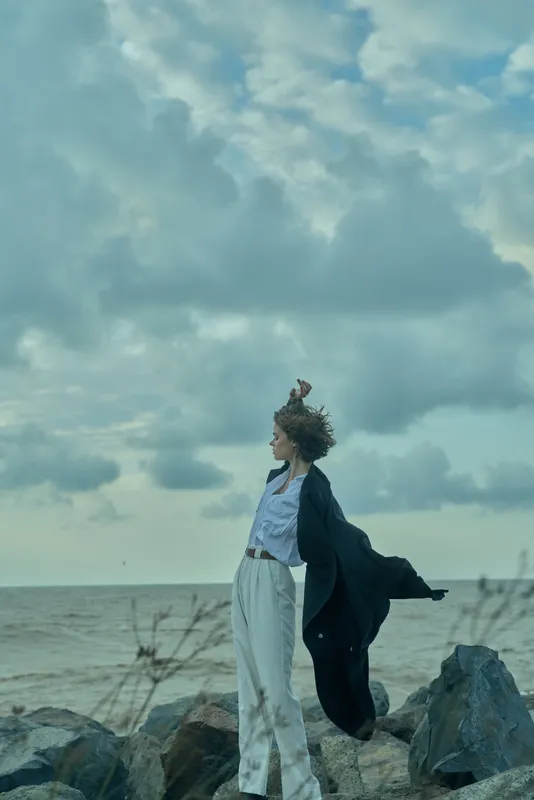 Windy beach scene with a woman in a flowing coat by the sea, embracing the breeze, open horizon, calm mood, coastal style, motion and freedom in a serene landscape