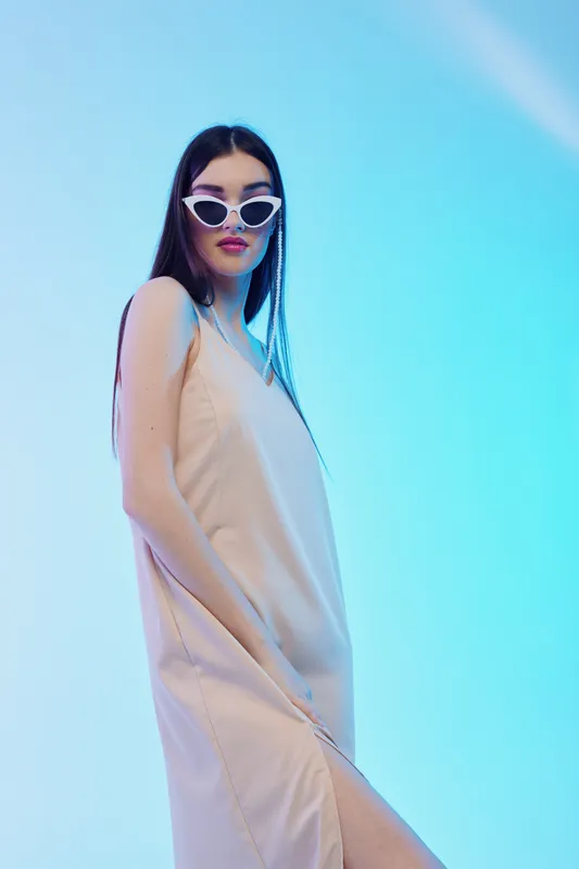 Woman beauty fashion model wearing white sunglasses and a beige dress posing confidently against blue background. Concept for style, elegance, and modern trends.